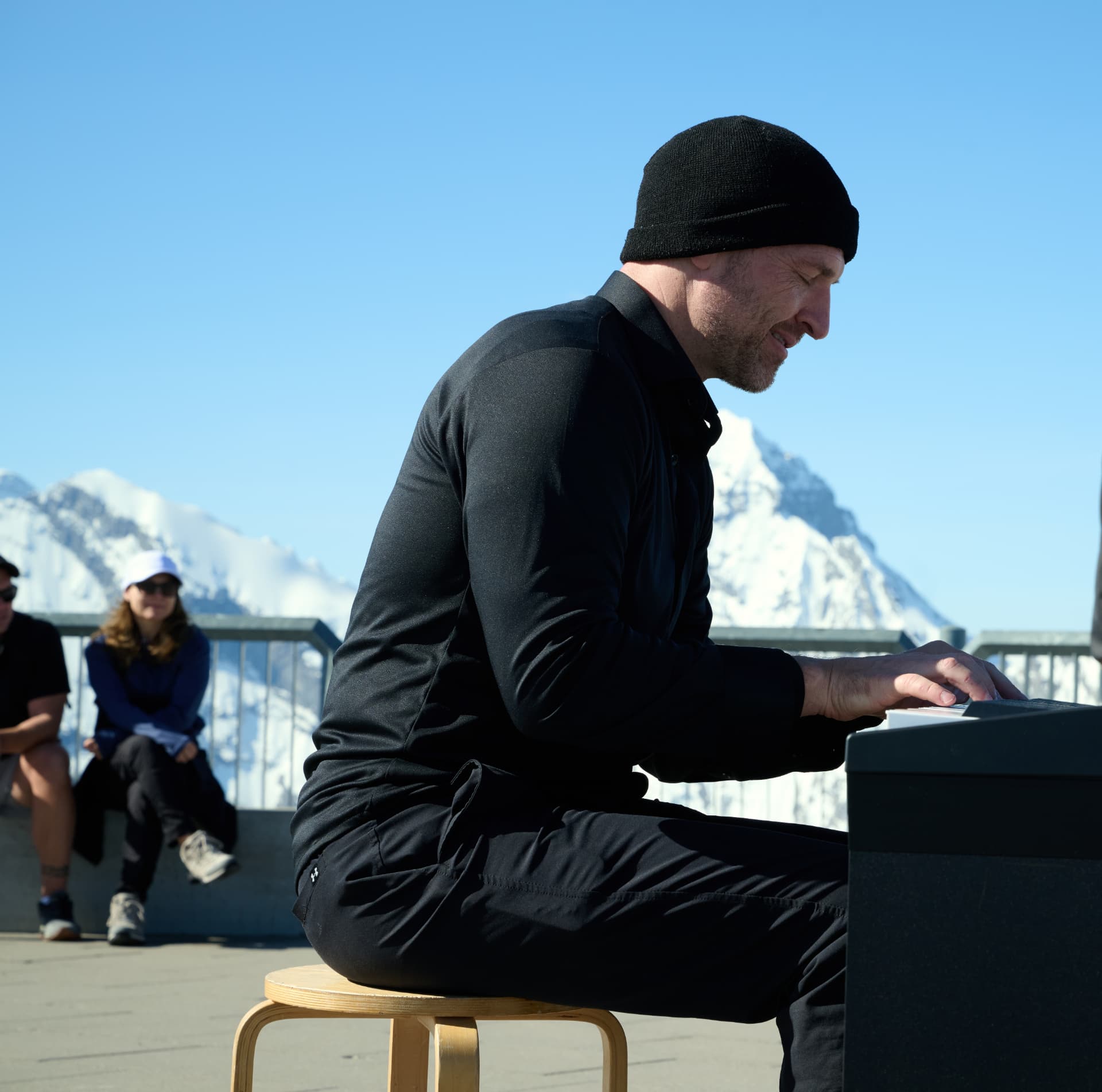 Matthew Mayer playing a portable keyboard on an alpine summit terrace, snowy peaks and a clear blue sky behind him, onlookers seated nearby