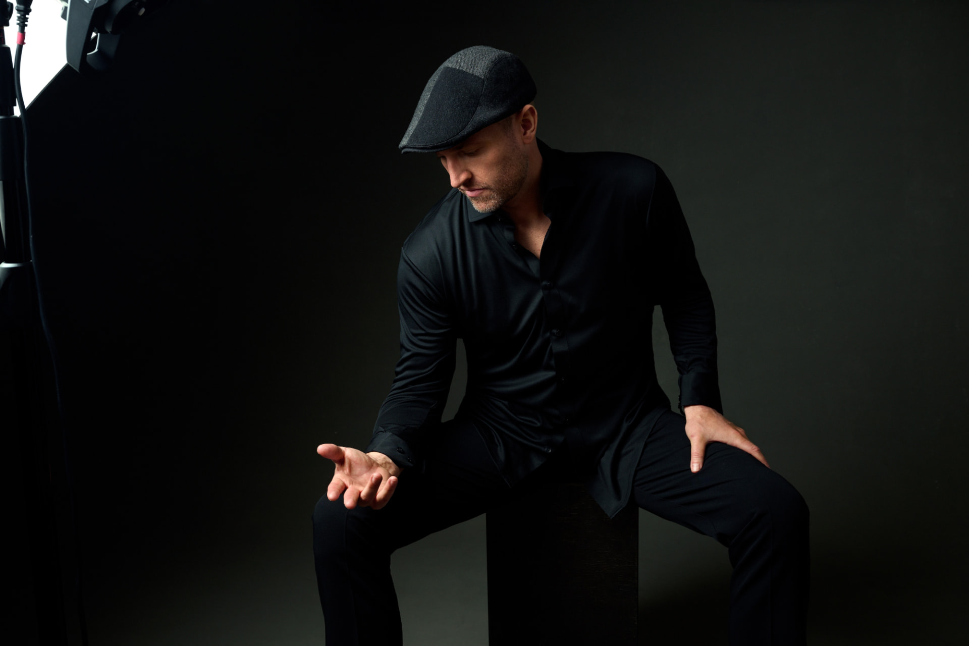 Studio portrait of Matthew Mayer seated in black with a flat cap, hands raised mid-gesture, against a dark charcoal backdrop