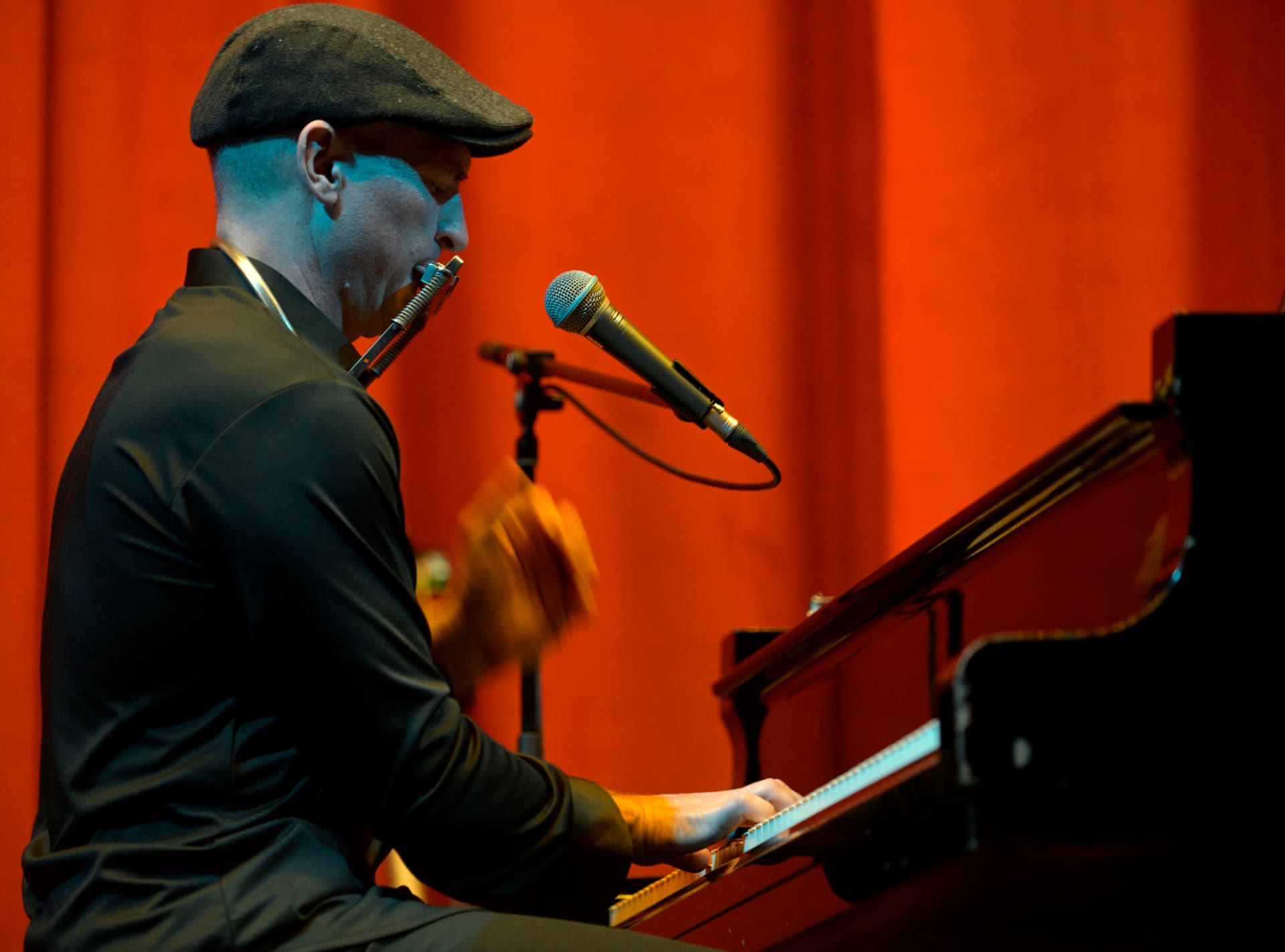 Matthew Mayer playing a grand piano and harmonica on stage before a red-lit curtain, Pancevo, Serbia