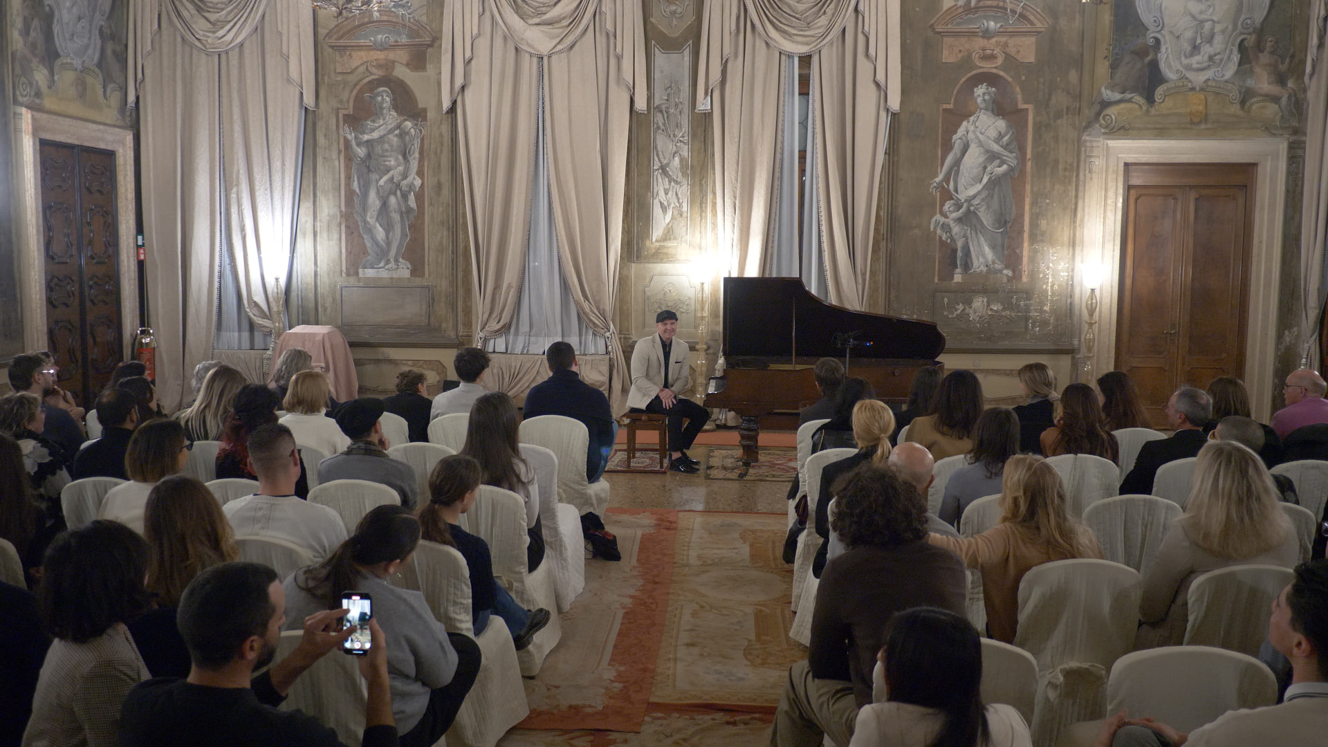 Matthew Mayer performing live at a grand piano before a seated audience in an ornate palazzo hall in Venice, Italy